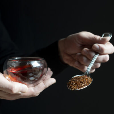 Una mano regge la tazza con il rooibos e l'altra mano un cucchiaio di foglie