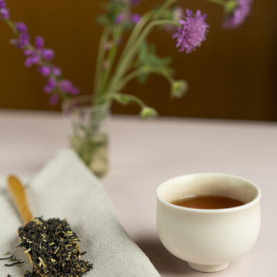 Una paletta di legno colma di foglie di tè e una tazza di tè. Sullo sfondo fiori di campo