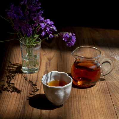 Una tazzina e una brocca di tè, sullo sfondo un bicchiere con fiori di campo