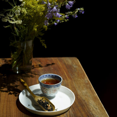 Foglie di tè su un cucchiaio di legno, una tazzina di tè, fiori recisi di campo sullo sfondo