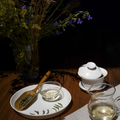 Un piattino bianco con le foglie asciutte e quelle bagnate, una brocca e una tazzina di tè, una gaiwan e sullo sfondo fiori di campo in un bicchiere