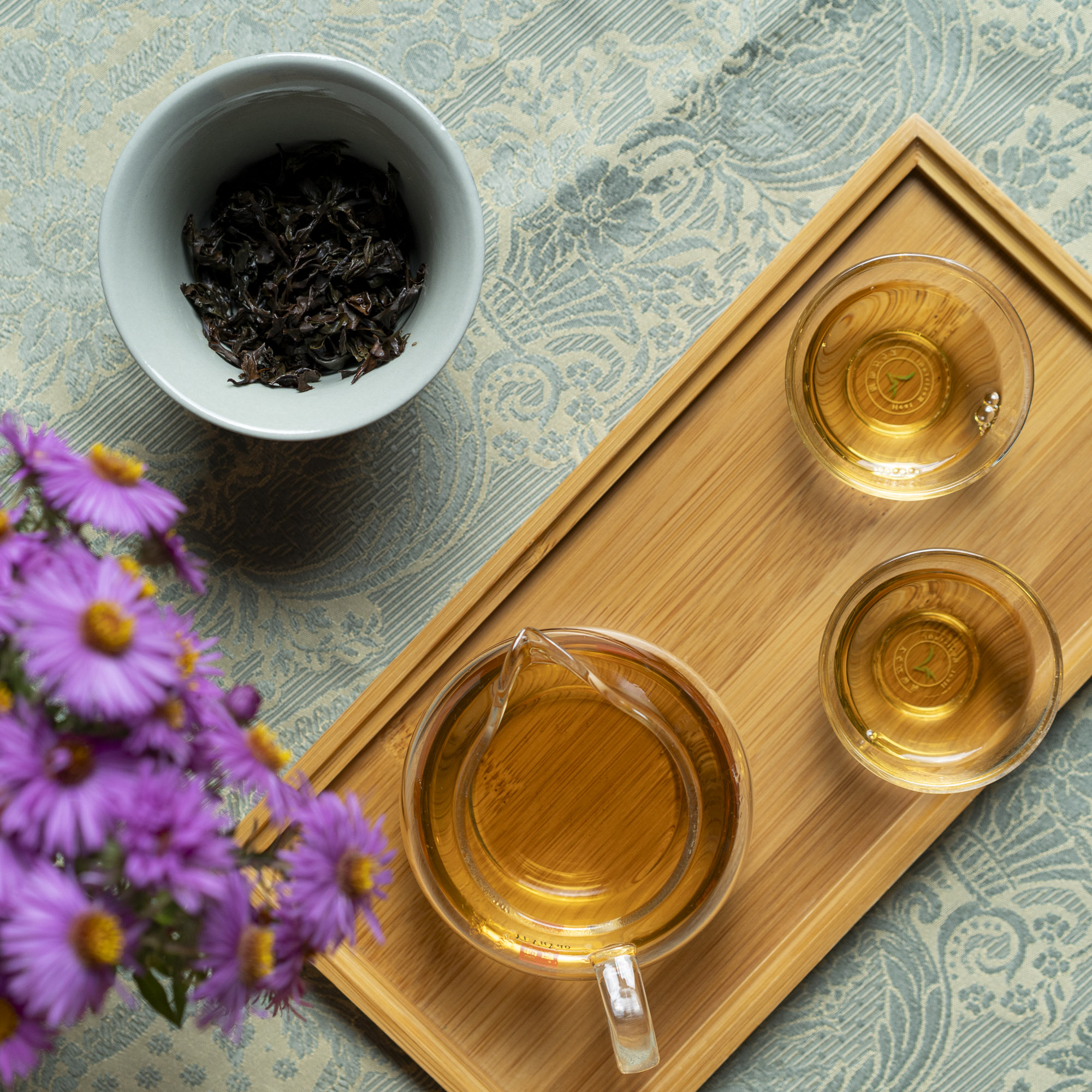 Due tazzine e una brocca di tè, una gaiwan aperta con le foglie bagnate e un mazzolino di settembrini viola.