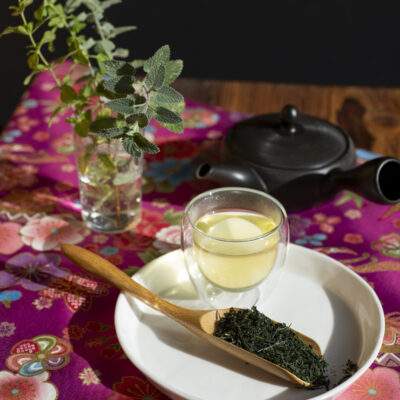 Foglie di Sencha, una tazza di tè e sullo sfondo la Kyusu nera e un vasetto con rametti di erbacee autunnali