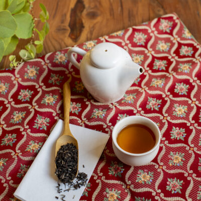 Un cucchiaio di legno con le foglie asciutte, una tazzina colma di tè e una piccola teiera bianca