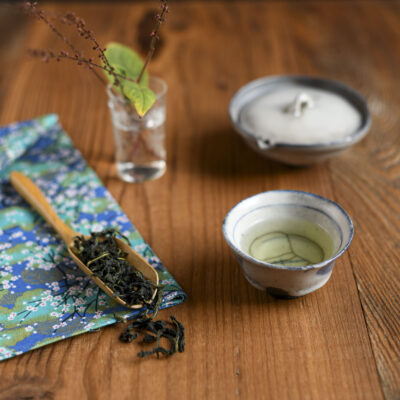 Un cucchiaio di tè con le foglie asciutte, una tazzina con il tè, una shiboridashi e un bicchiere con un delicato fiore autunnale