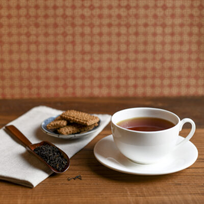 Una tazza bianca con il tè pronto, accompagnata da un piattino di biscotti e da un cucchiaio di legno colmo di foglie di tè asciutte