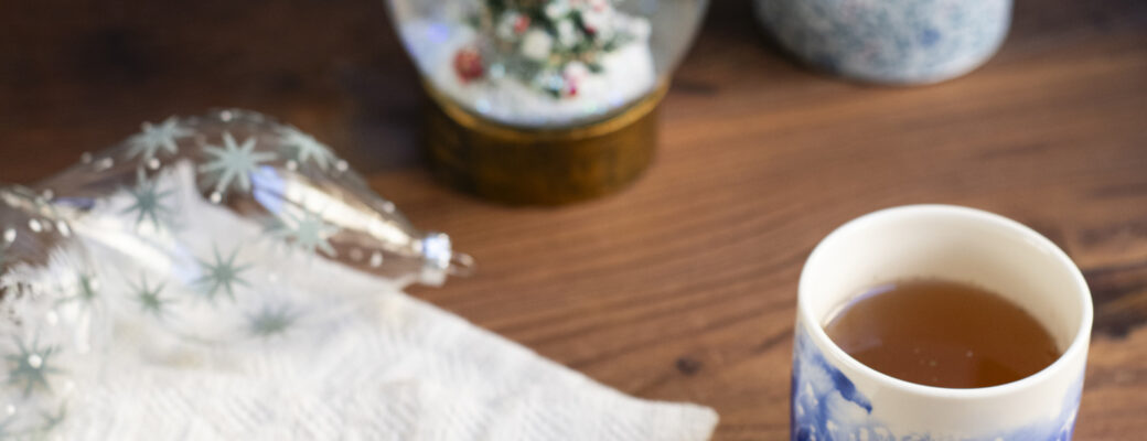 Un piattino con alcune foglie di tisana di Natale. Sullo sfondo un bicchiere con decori blu, una scatola porta tè, delle palline di Natale di vetro e una palla di vetro innevata...