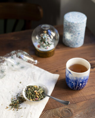 Un piattino con alcune foglie di tisana di Natale. Sullo sfondo un bicchiere con decori blu, una scatola porta tè, delle palline di Natale di vetro e una palla di vetro innevata...