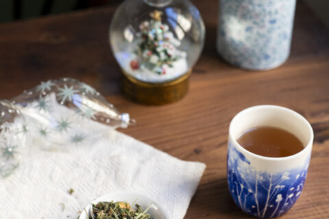 Un piattino con alcune foglie di tisana di Natale. Sullo sfondo un bicchiere con decori blu, una scatola porta tè, delle palline di Natale di vetro e una palla di vetro innevata...